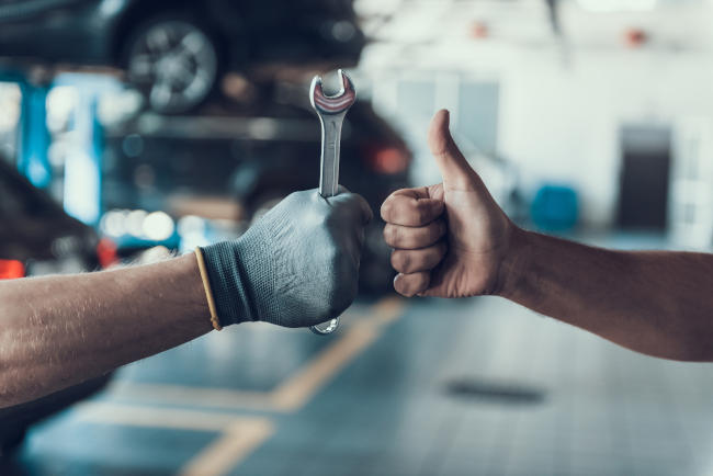 An image of a gloved hand holding a wrench, fist-bumping a second hand with no glove, giving a "thumbs up." In the background is an automotive service garage. 