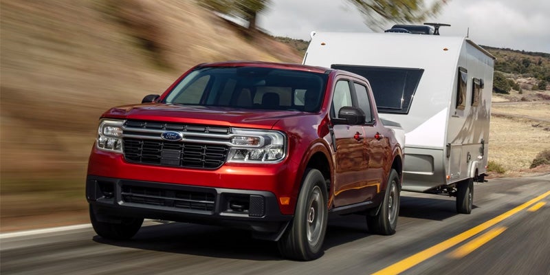 A red 2024 Ford Maverick pulling a trailer down a mountain road. 