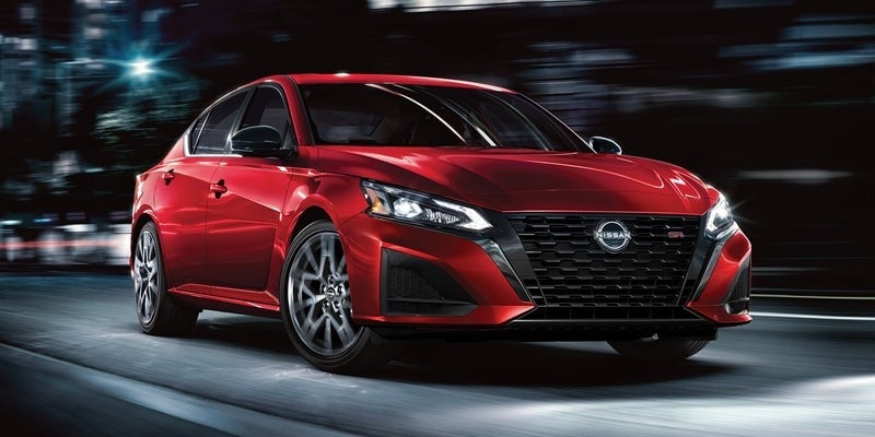 A metallic red Nissan Altima driving down a city street at night. The background is blurred to give the indication of speed. 