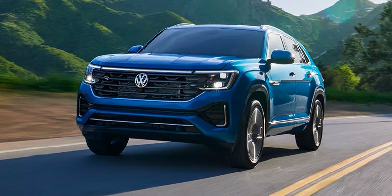A metallic blue VW Atlas Cross Sport driving down a highway backed by scenic mountains.